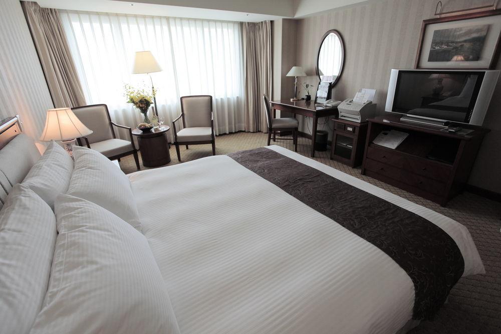 Evergreen Laurel Hotel - Тайджун Екстериор снимка The photo shows a well-furnished hotel room. In the foreground, there is a large bed with white bedding and a dark-colored throw at the foot. Beside the bed, there is a bedside lamp. 

In the background, natural light filters through large windows co