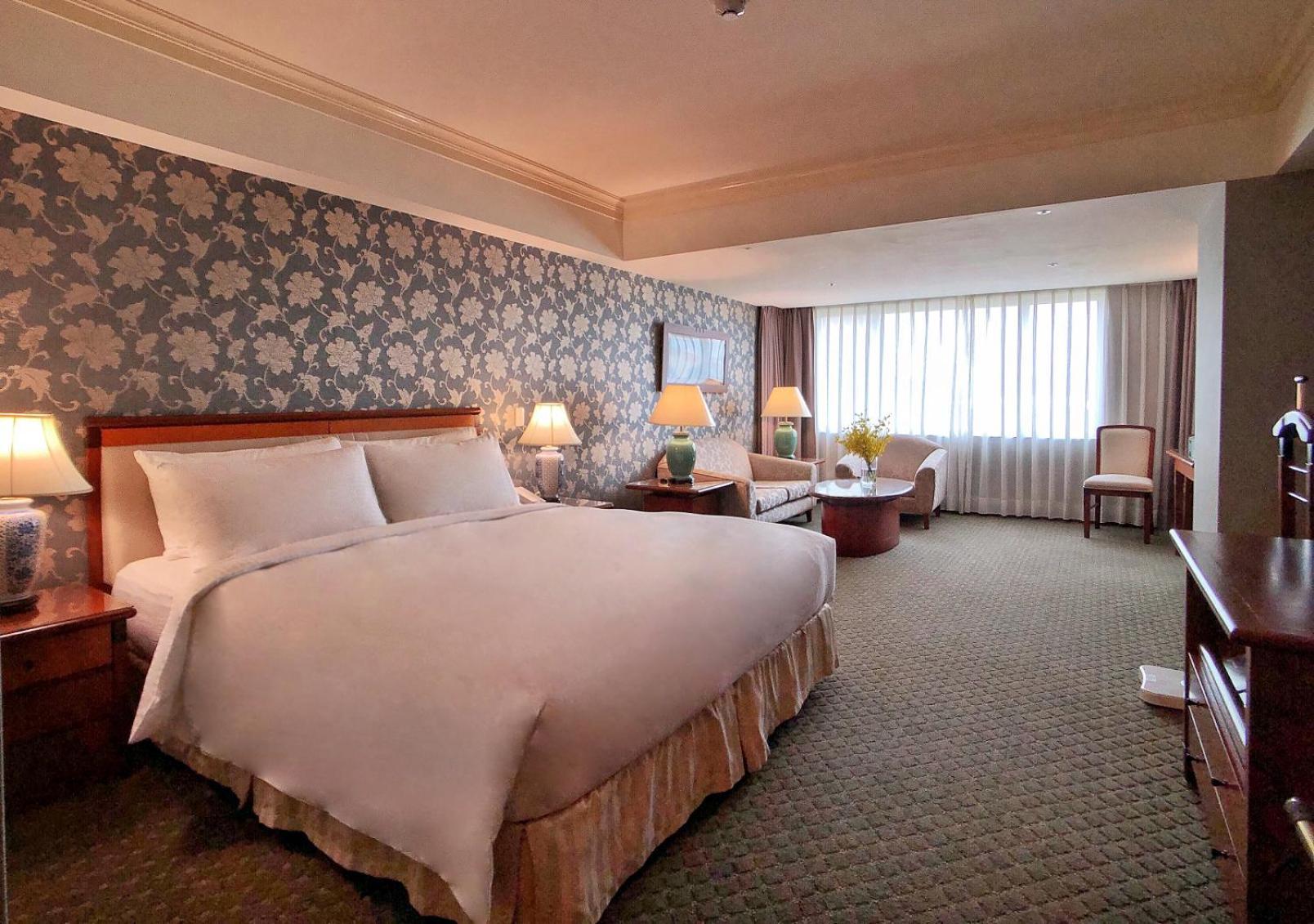 Evergreen Laurel Hotel - Тайджун Екстериор снимка The photo shows a hotel room with a spacious and comfortable layout. It features a large bed with white bedding, placed against a decorative wallpapered wall. On either side of the bed, there are lamps providing soft lighting. In addition to the bed,