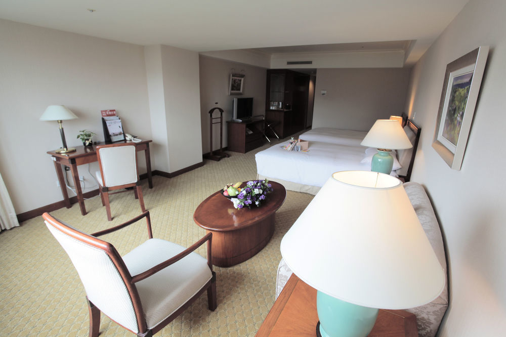 Evergreen Laurel Hotel - Тайджун Екстериор снимка The photo shows an interior view of a hotel room. It features two beds, a coffee table with a small decorative arrangement of flowers, and a comfortable chair. There's a desk and a lamp, along with another larger lamp on the table. In the background,