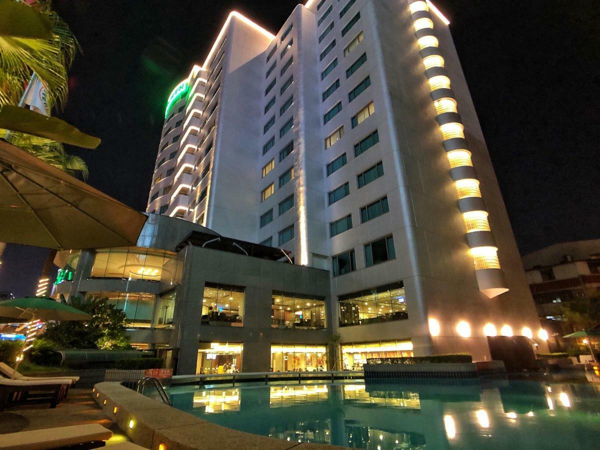 Evergreen Laurel Hotel - Тайджун Екстериор снимка The photo shows a tall hotel building at night, illuminated by lights. The architecture appears modern, with several floors and large windows. In the foreground, there is a swimming pool that reflects the lights from the building, creating a serene a