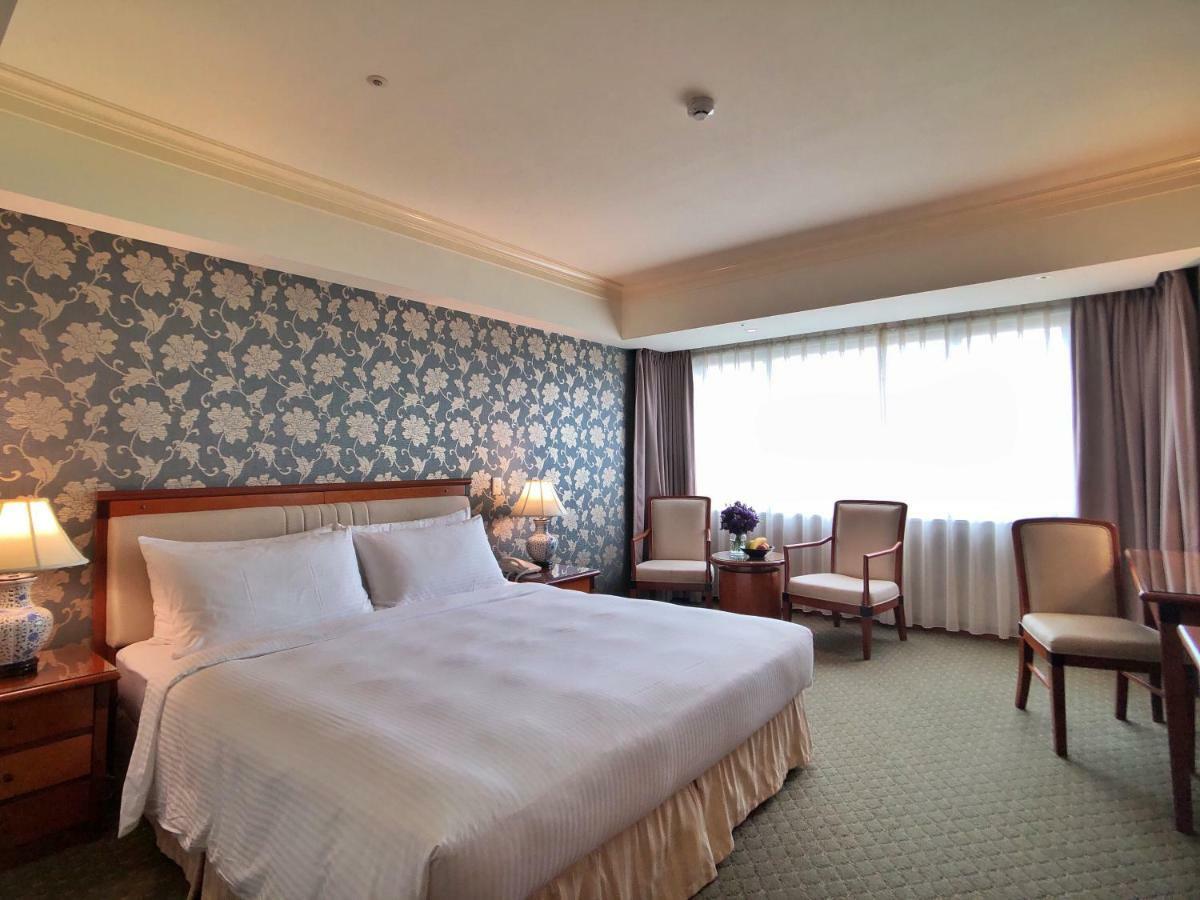 Evergreen Laurel Hotel - Тайджун Екстериор снимка The photo shows a hotel room that features a large bed with a white bedspread positioned against a decorative floral wallpaper. There are two bedside lamps on wooden nightstands flanking the bed. To the side of the room, there are two chairs and a sm