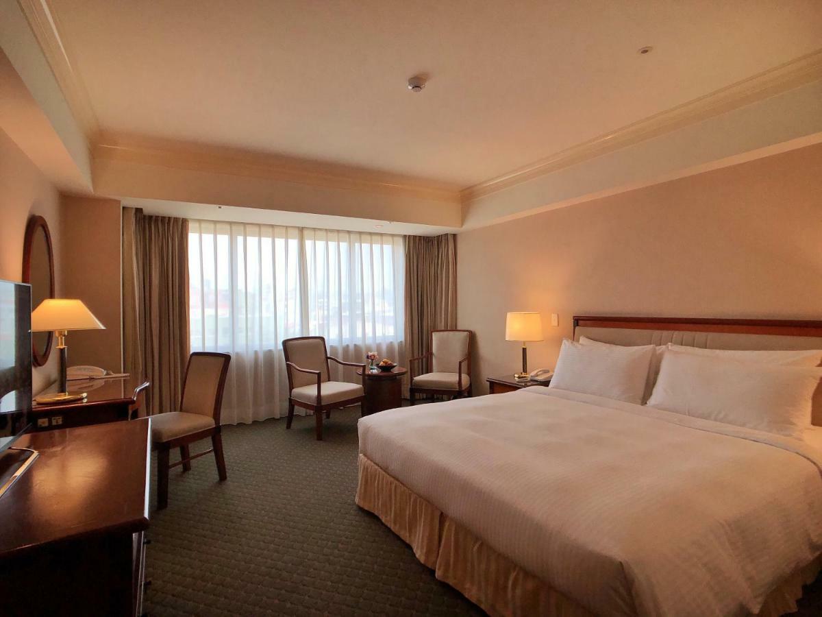 Evergreen Laurel Hotel - Тайджун Екстериор снимка The photo shows a hotel room featuring a large bed with white linens, a wooden bedside table with a lamp, and a flat-screen television. There are two chairs with a small table next to them positioned by a large window, which has sheer curtains lettin