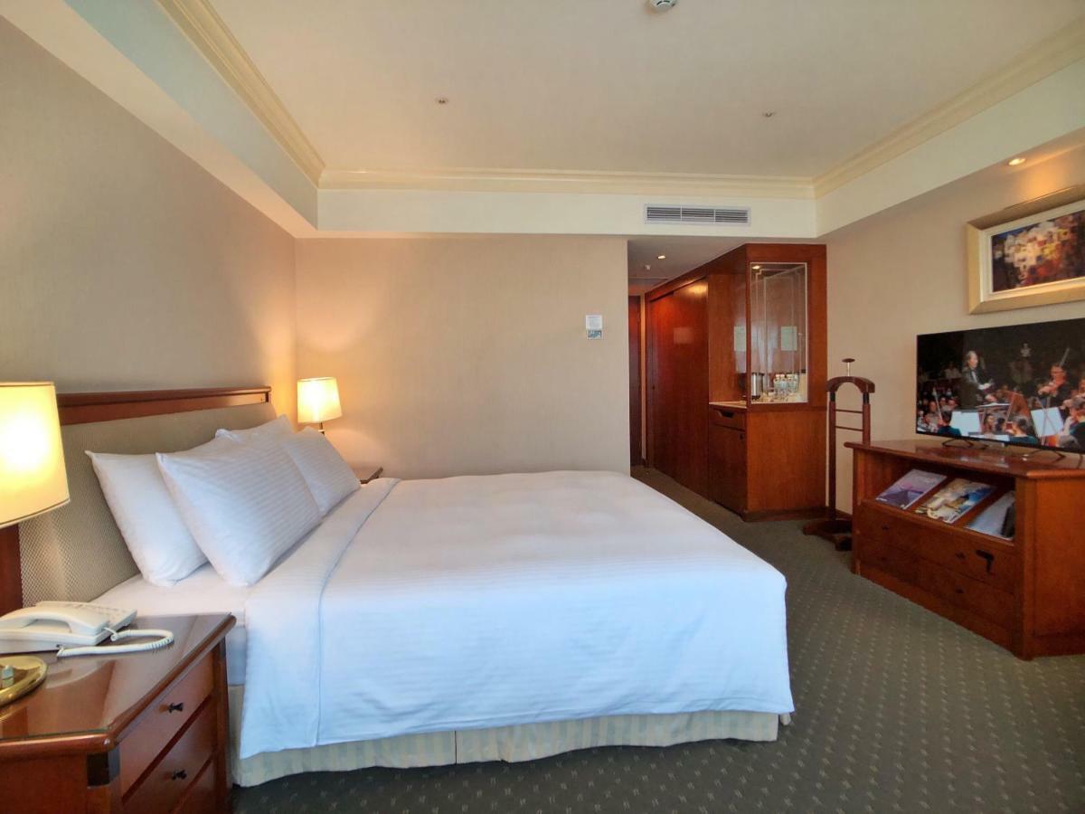 Evergreen Laurel Hotel - Тайджун Екстериор снимка The photo shows a hotel room featuring a comfortable bed with white linens and pillows, positioned against a light-colored wall. There is a wooden bedside table with a lamp and an open book next to the bed. On the opposite side, there’s a large flat-