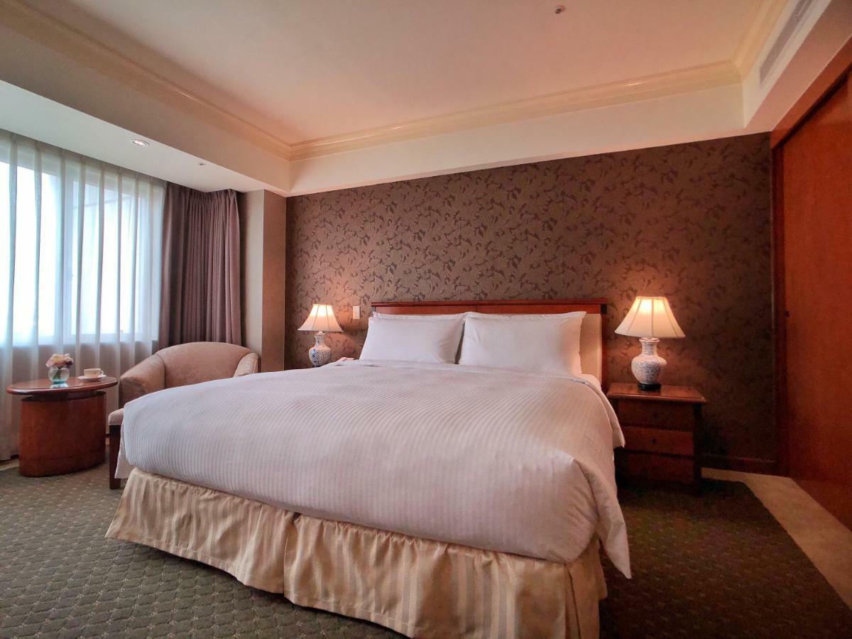 Evergreen Laurel Hotel - Тайджун Екстериор снимка The photo shows a neatly arranged hotel room featuring a large bed with white linens and a light-colored bed skirt. On either side of the bed, there are small wooden nightstands with lamps. To the left of the bed, there's a comfortable chair and a ro